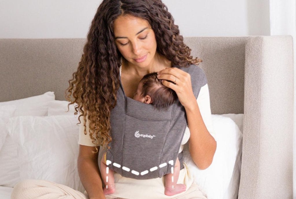 Woman holds baby in an Ergo Baby Carrier touching its head. A dotted line across the legs and bum shows "M" positioning