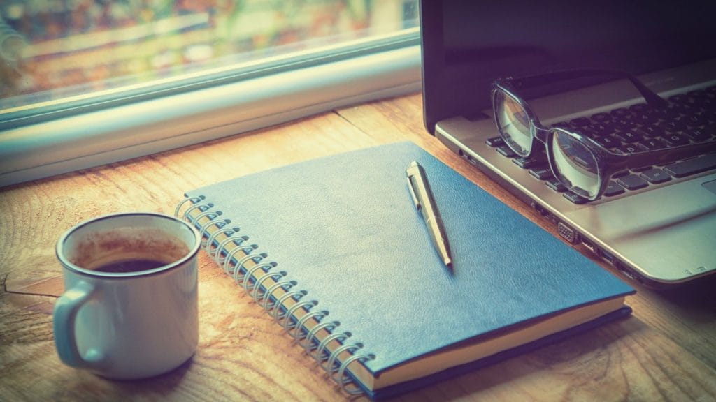 a cup of coffee, blue notebook with a pen, and a laptop sit on a desk.