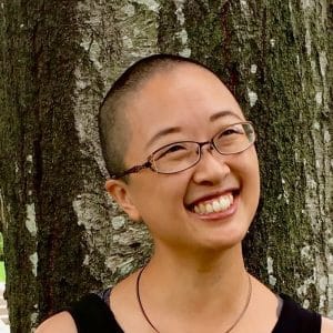 Color photo of Angelique Geehan's head and shoulders in front of a mottled gray tree trunk. She is a medium-toned East Asian person with a buzzed head of black hair, wearing glasses, smiling and looking off to the right, with her head tilted slightly. Also wearing a black wire necklace with silver metal rings looped onto it and a black tank dress.
