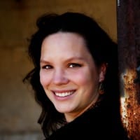 Color photo of Kristi Hayes-Devlin with her head turned to look directly at the camera. She is a white-skinned woman with brown eyes. Her dark brown hair is loose. She is leaning against a rusted metal doorframe and smiling broadly.