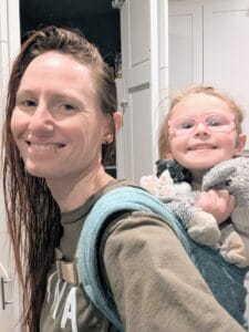 Sarah Zinn smiles at the camera. Her toddler is on her back in a baby carrier also smiling broadly. 