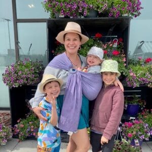 Allie Dennis and her three children smile directly at the camera. They are all wearing hats and they stand in front of a display of flowers. She is wearing her baby in a lavender ring sling. 