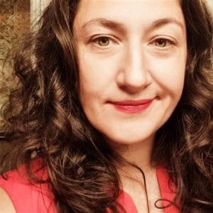 In this closeup, Geta smirks at the camera. Her brown hair is in waves, her eyes are bright, and she is wearing lipstick that matches her shirt. 