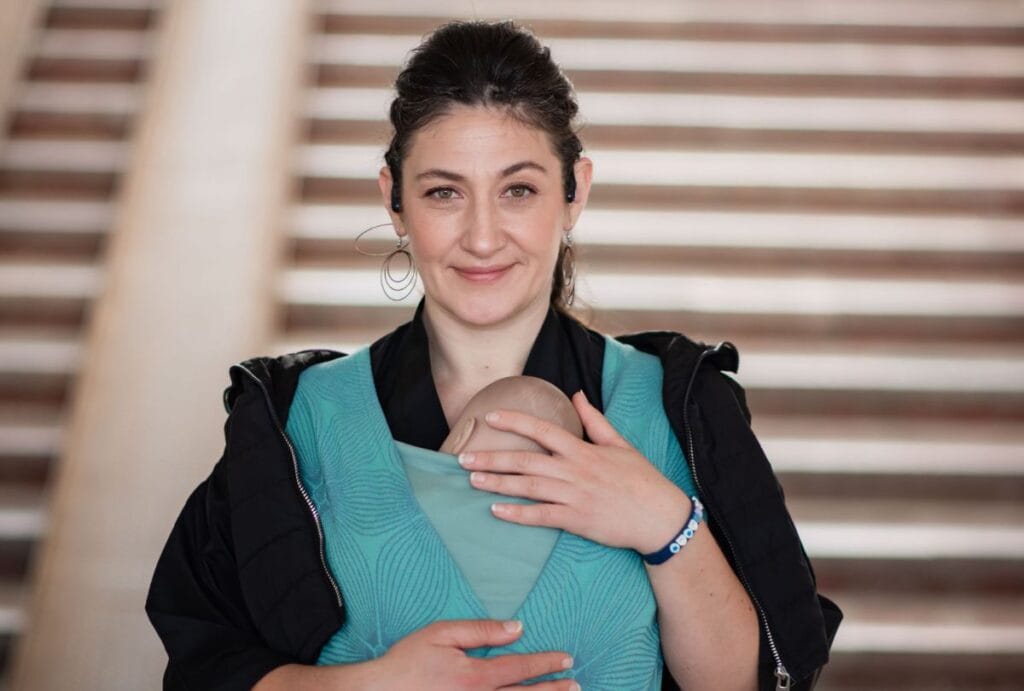 A doll is wrapped in a teal wrap and the adult is smiling at the camera. They're standing in front of a staircase.