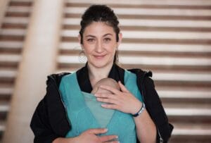 A doll is wrapped in a teal wrap and the adult is smiling at the camera. They're standing in front of a staircase.