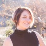 Allie Vella stands in front of a tree. She smiles directly into the camera. She has dark hair and wears a black shirt.