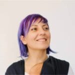 Diana Mateescu stands in front of a white wall. She is looking to her left and smiling. Her hair is purple and her shirt is black.