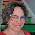 Kristi Hayes-Devlin smiles at the camera. She has short, curly salt and pepper hair and is wearing iridescent glasses. She sits on of rainbow staircase.